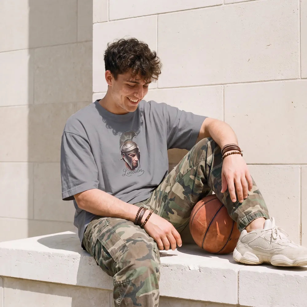 Man sitting on a pale stone ledge beside a basketball, in an oversized grey tee printed with Abraham Lincoln in a crested helmet