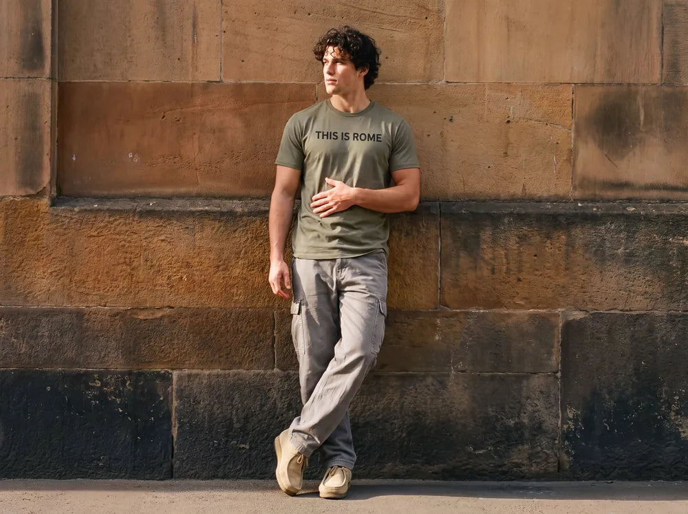 Man leaning against a sunlit sandstone wall in an olive tee printed THIS IS ROME
